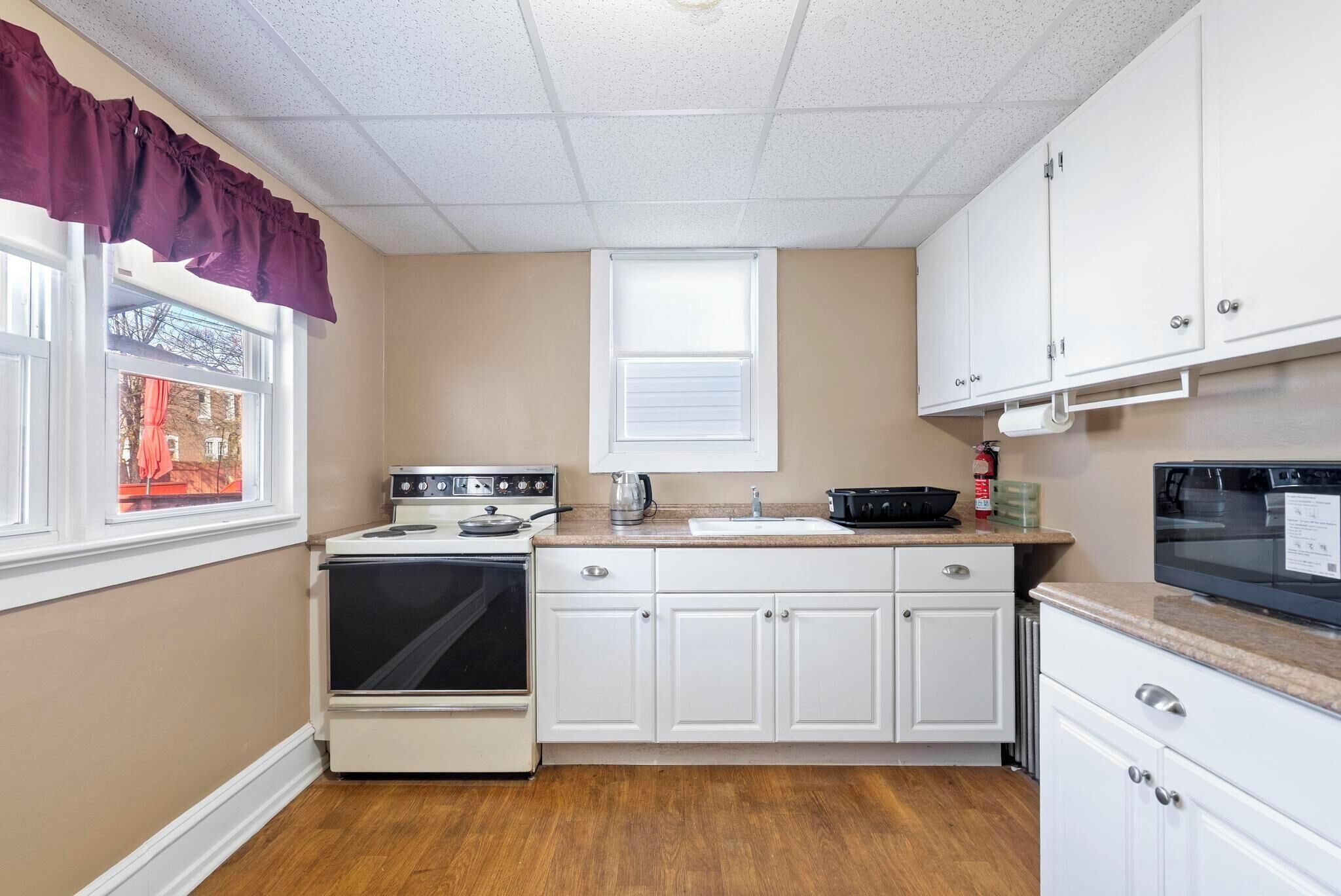 A compact kitchen with white cabinets, a stove, a microwave, and a window with a purple valance.