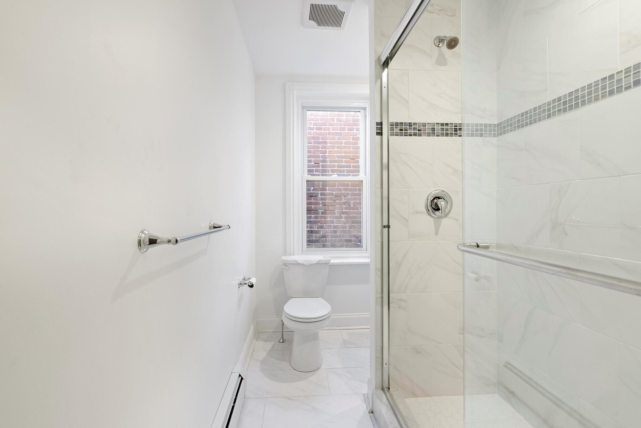 A modern bathroom with a glass shower enclosure, a toilet, and a window with a brick wall view.