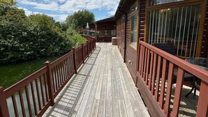A wooden deck leading to a cabin with large windows, a view of the sky, and a wooden railing. - Kingfisher Cosy Lakeside Lodge (Carnforth)