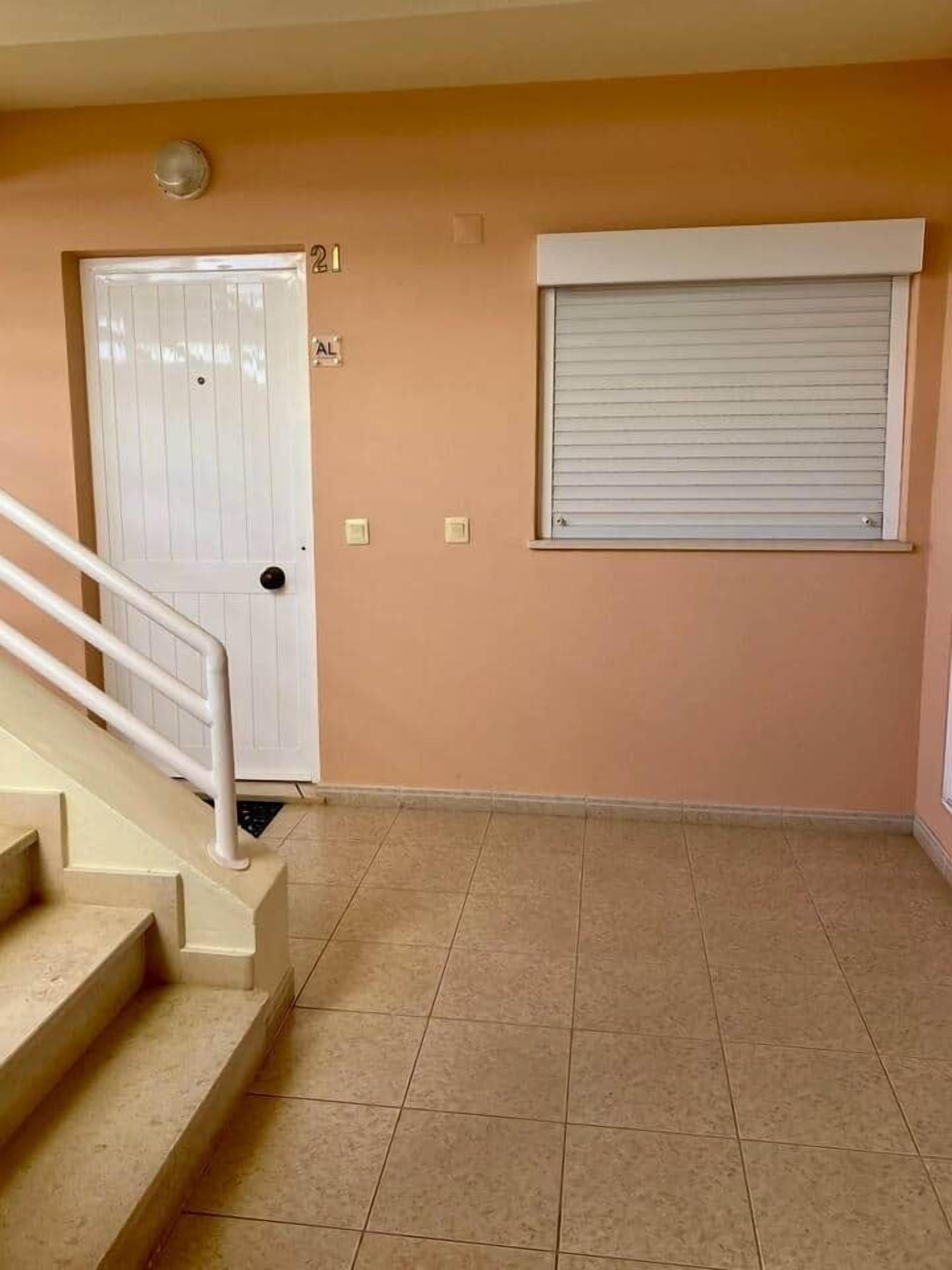 Un pasillo con una puerta blanca marcada con el número 21 y una ventana con una persiana cerrada.