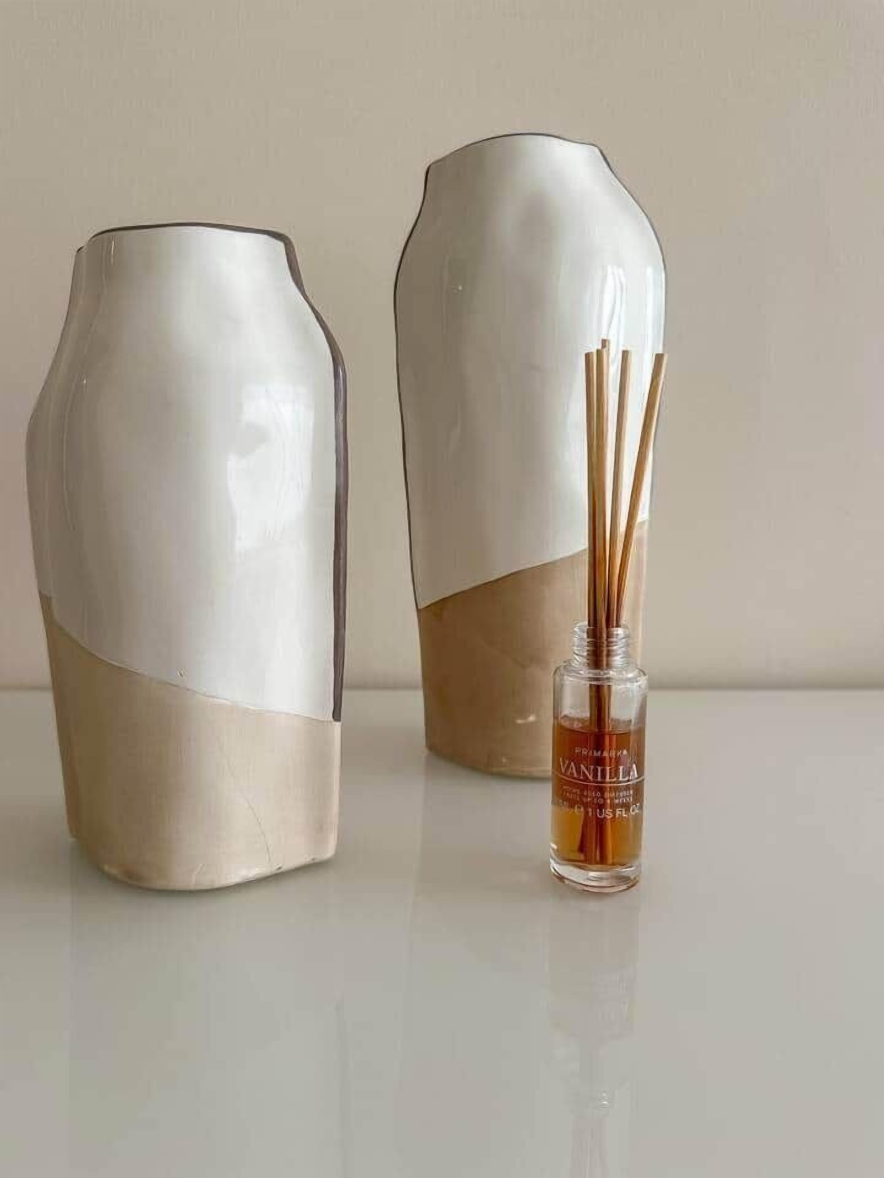 Deux vases en céramique aux tons beige et blanc, l’un avec un col plus étroit et l’autre avec une base plus large, et une petite bouteille de diffuseur d’ambiance à bâtonnets parfumés à la vanille placée devant eux.