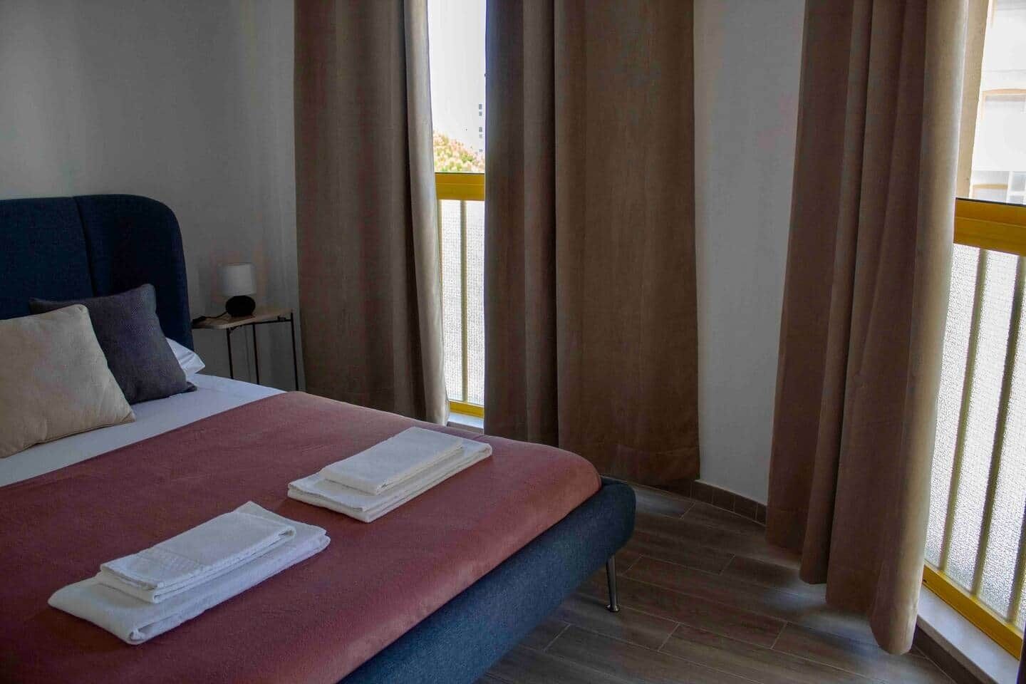 Una habitació d'hotel amb llit, cortines, una tauleta de nit i una finestra amb vista.