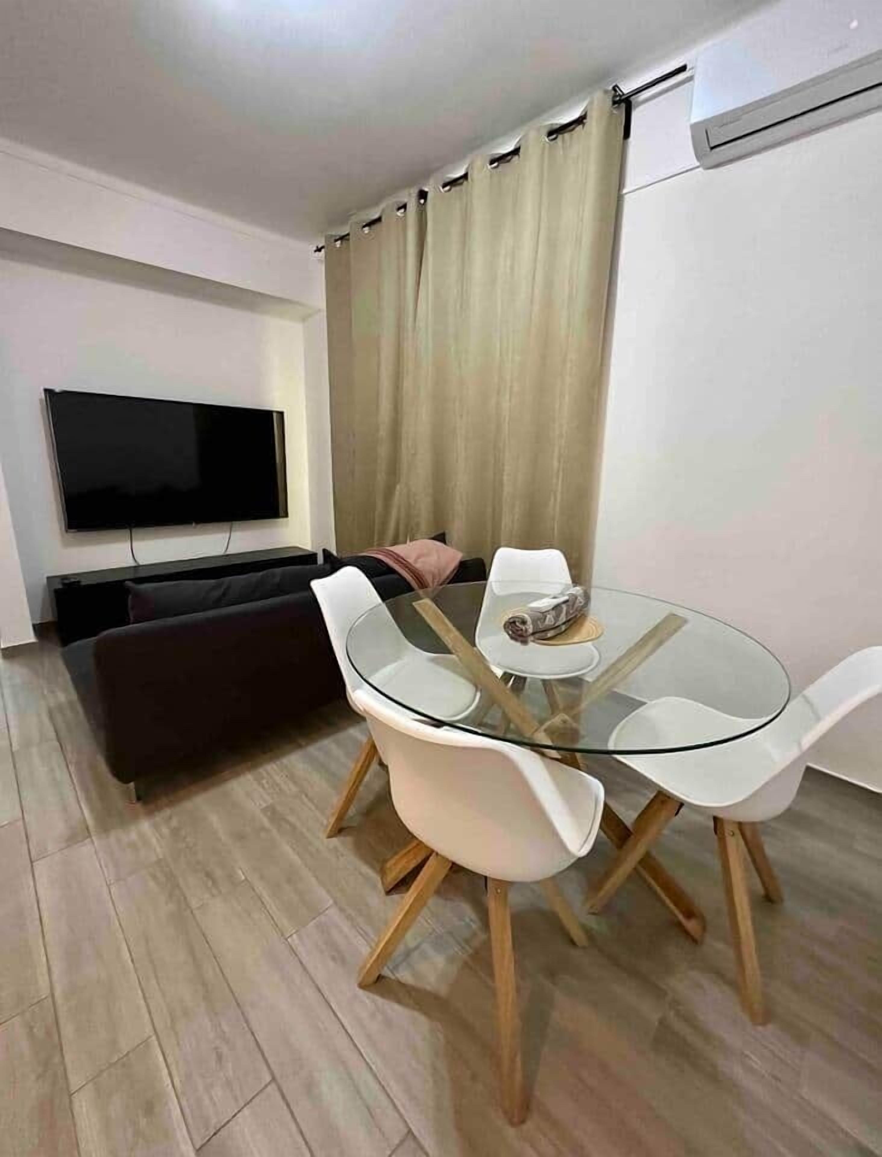 A modern living room with a flat-screen TV, a sofa, a glass dining table with chairs, and a large window with curtains.
