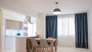 A modern kitchen with white cabinets, a dining area with a wooden table and chairs, and a large window with blue curtains. - Seaside Inspire Wilderness (Quarteira)