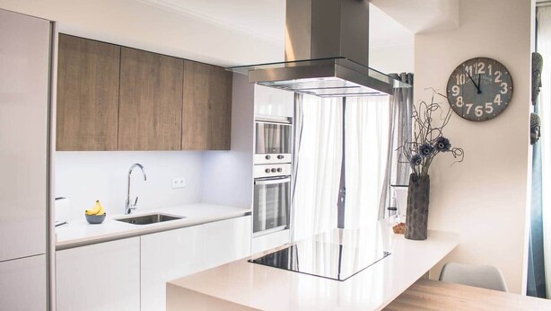 A modern kitchen with white cabinetry, a stainless steel range hood, and a wall-mounted clock. - Seaside Inspire Wilderness (Quarteira)