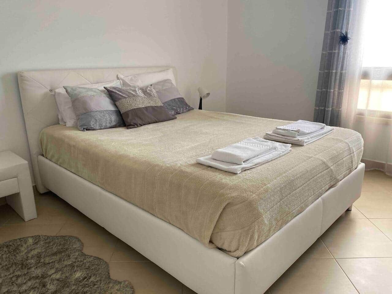 A neatly made bed with a beige bedspread and pillows, a bedside table with a lamp, and a window with curtains.