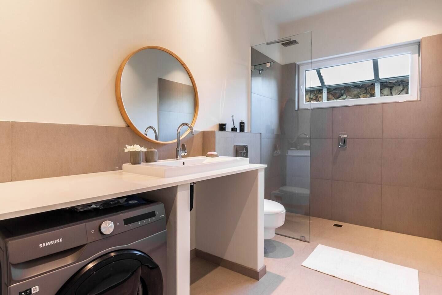 Un baño moderno con un espejo grande, una lavadora Samsung, un inodoro y una zona de ducha.