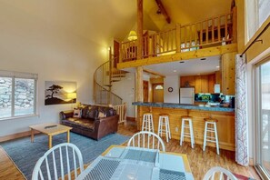 A spacious living area with a high wooden ceiling, a cozy seating area, a kitchen with wooden cabinets, and a visible staircase leading to an upper level.