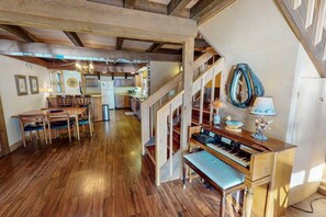 Una habitación amplia con suelo de madera, una zona de comedor con sillas, un piano y una escalera.