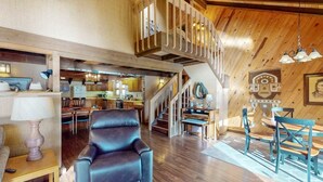 A spacious living area with a high ceiling, wooden beams, a staircase, and a dining area with blue chairs.