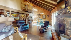 A spacious living room with a fireplace, wooden beams, and a staircase leading to an upper level.