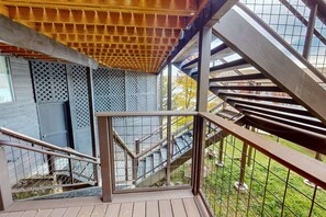 Une terrasse en bois avec un escalier en métal menant à une zone abritée d’où l’on peut admirer un bâtiment et un espace verdoyant.
