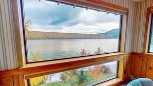 A room with a large window overlooking a lake, a chair, and wooden paneling. - The Treehouse lakefront– scenic family getaway (Joseph)