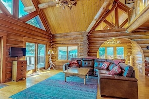 A log cabin interior with a wooden ceiling, a large window, a television, a sofa, and a patterned rug.