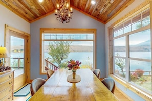 Comedor con mesa de madera, sillas, candelabro y ventanas amplias con vista al lago.