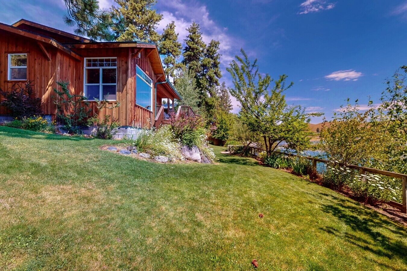 Una cabaña de madera con un ventanal grande y una terraza, rodeada de un césped cuidado y árboles.
