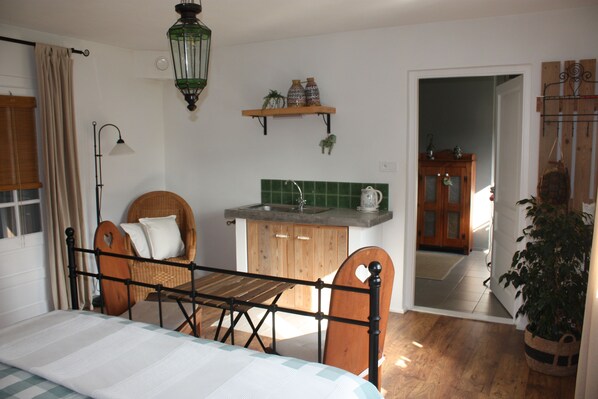 Un dormitorio con cama, una silla de madera, un pequeño rincón de cocina y una puerta que da a otra habitación.