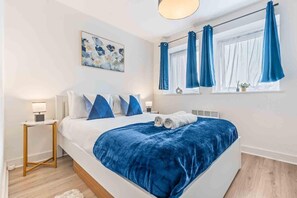 A modern bedroom with a large bed, blue bedding, a painting on the wall, and a window with curtains. - Couples- Business- Sleeps 2- Wifi- Pets- Parking (Coventry)