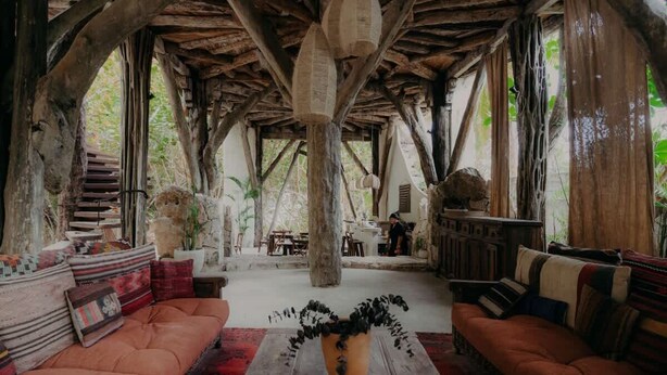 Un interior rústico con vigas de madera, una zona de estar central con sofás, una mesa baja y una lámpara colgante.