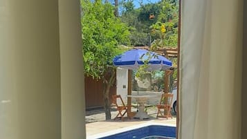Une vue à travers une fenêtre donnant sur une piscine, des meubles d’extérieur et de la verdure.