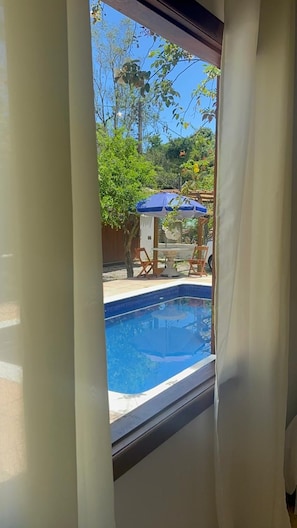 A view through a window with a pool, outdoor furniture, and greenery. - Pousada ViniCarmel (Porto Seguro)