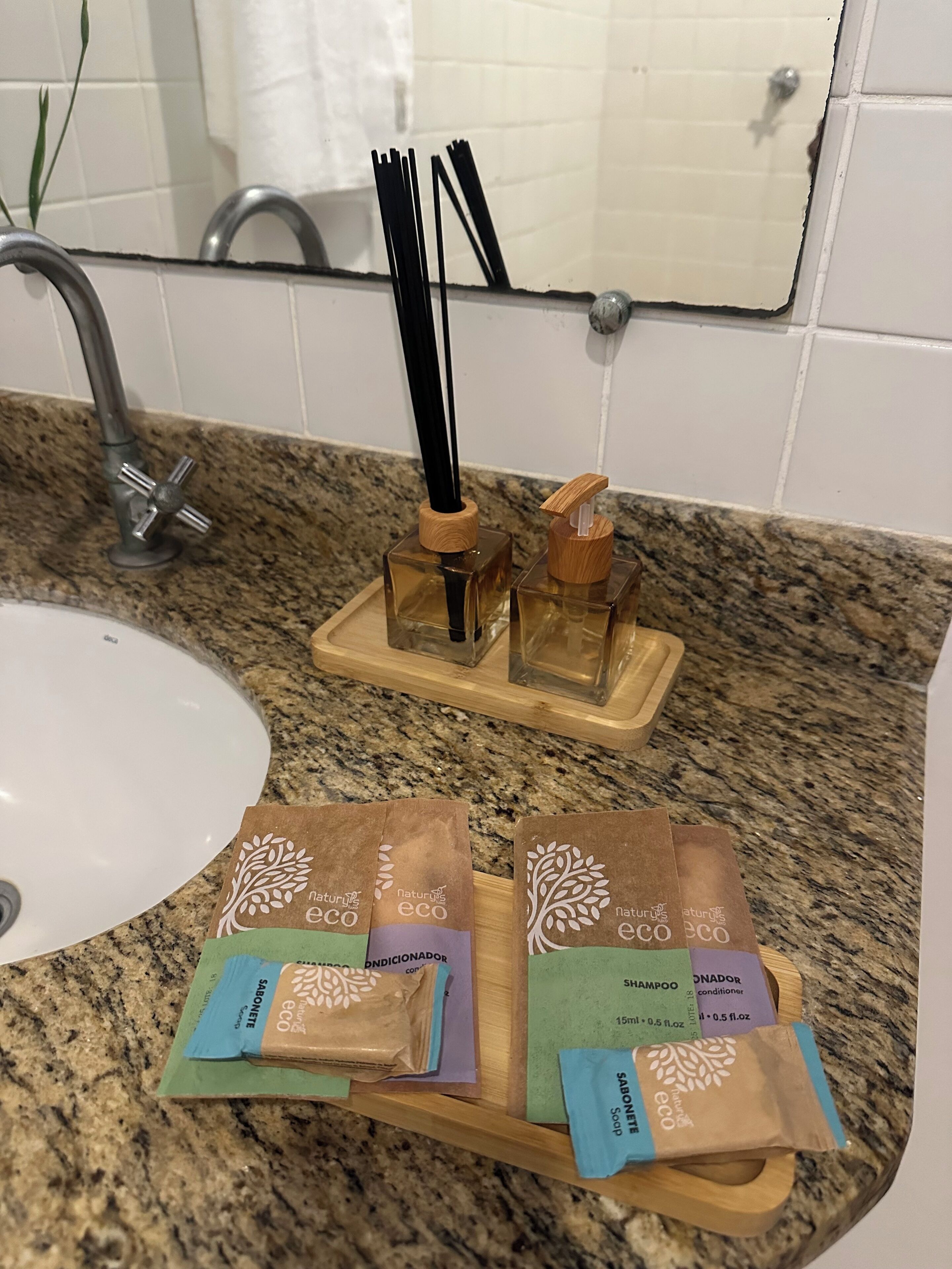 Baño con encimera de granito, lavamanos y espejo. Hay dos soportes de madera con palitos negros y de madera, y tres bolsas de papel marrón con marca ecológica.