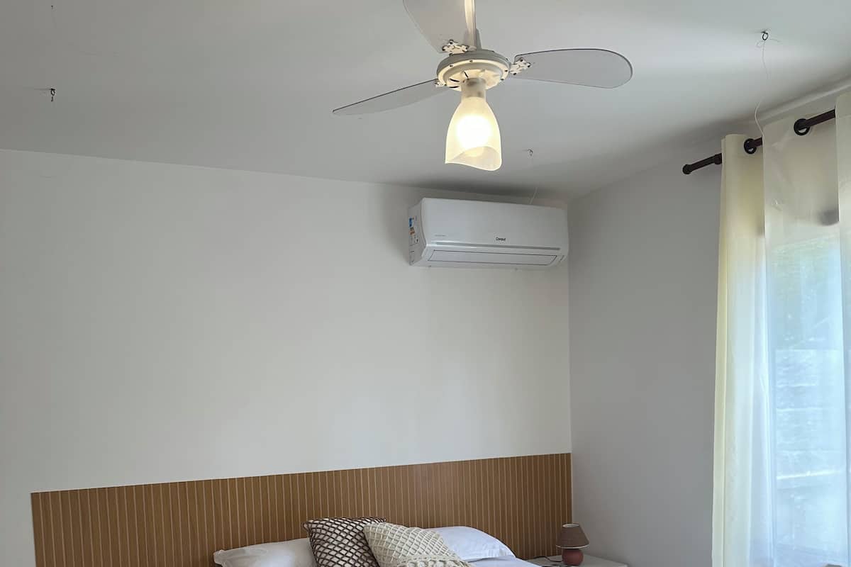 A hotel room with a bed, a nightstand, a ceiling fan, an air conditioning unit, and a window with curtains.