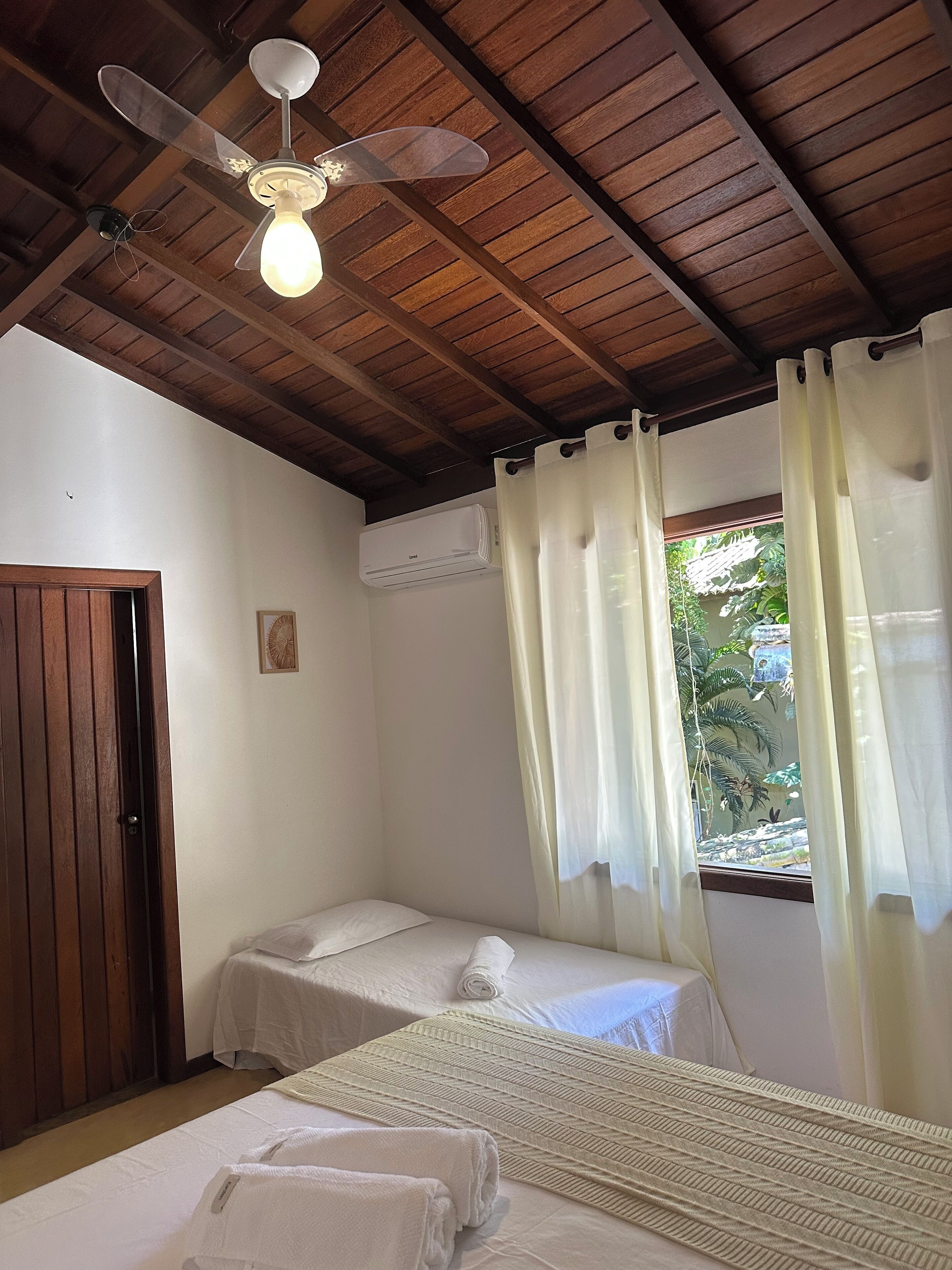 A bedroom with two single beds, a ceiling fan, an air conditioning unit, and a window with curtains.