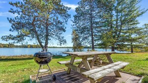 Una mesa de picnic con una parrilla, ubicada en un patio de ladrillo junto a un lago con árboles al fondo.