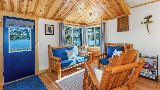 Interior de una cabaña de madera con un sofá azul, sillas de madera, una mesita y vistas al lago a través de grandes ventanales.