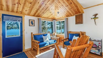 A wooden cabin interior with a blue sofa, wooden chairs, a small table, and a view of the lake through large windows.