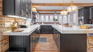 Een moderne keuken met donkere kastjes, witte aanrechtbladen en een centraal eiland. Er zijn hanglampen, een bakstenen muur en grote ramen.