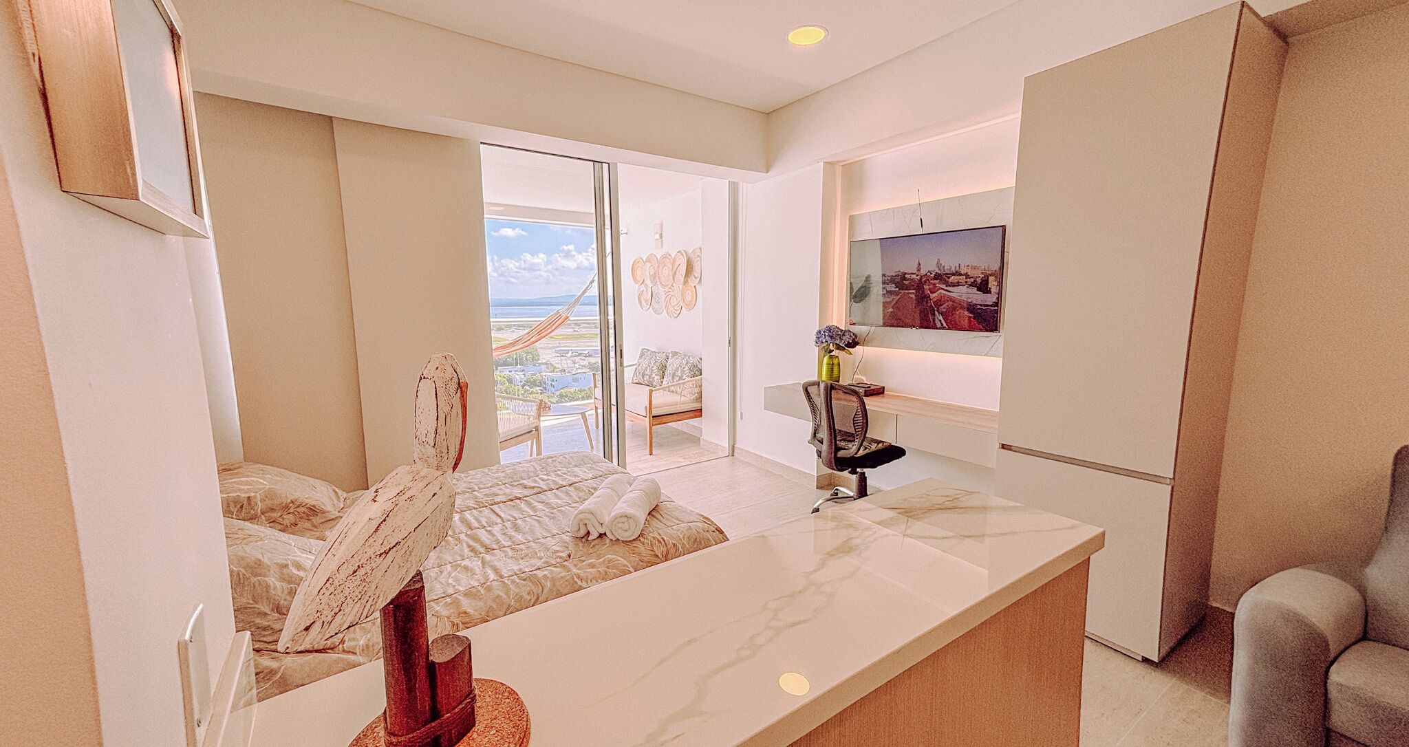 Habitación de hotel moderna con una cama grande, una zona de estar con una silla, un escritorio y un ventanal con vistas a la ciudad.