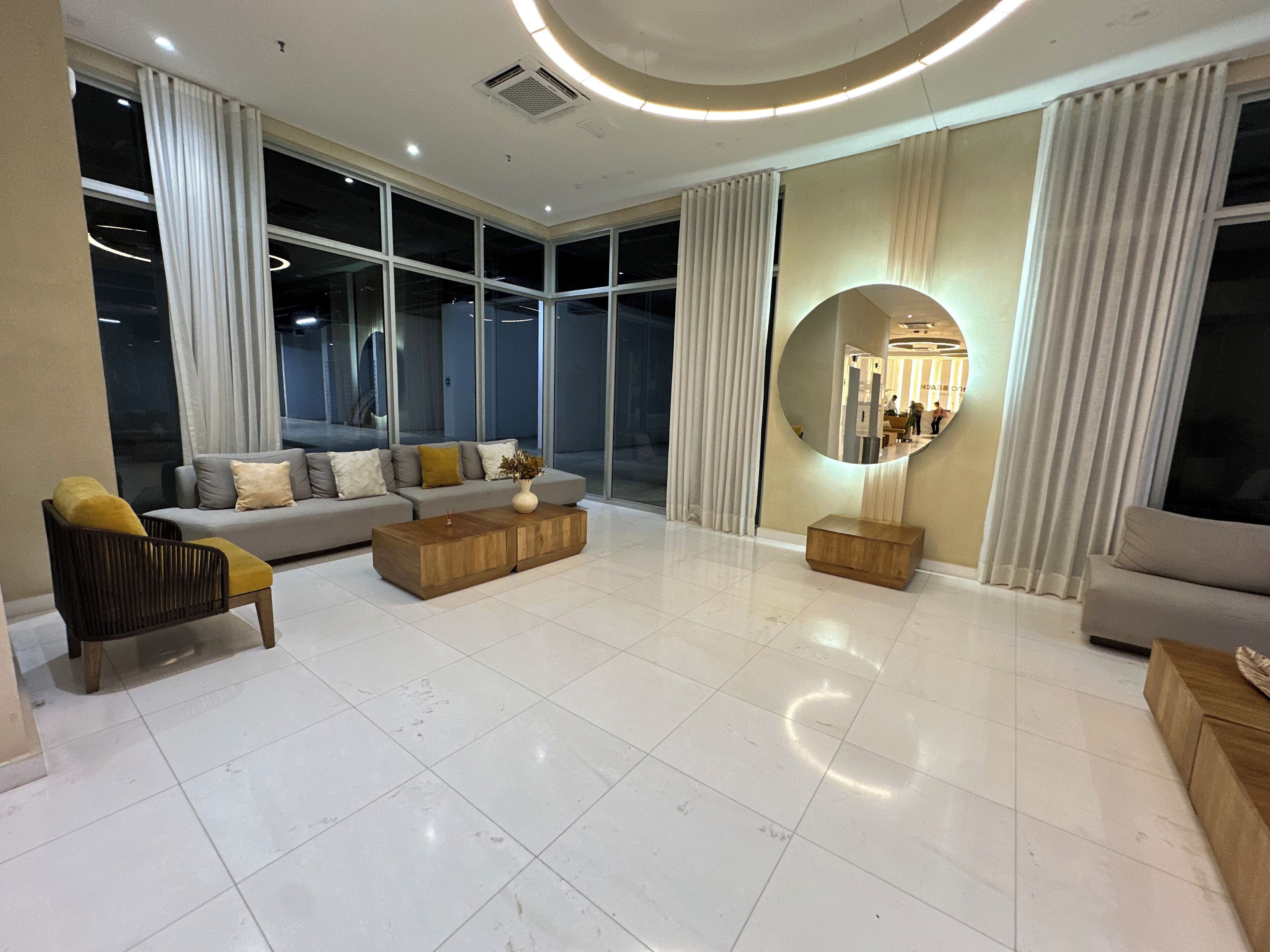 Una sala de estar moderna con un sofá, una mesa de centro y un sillón, ventanas amplias y un espejo redondo.