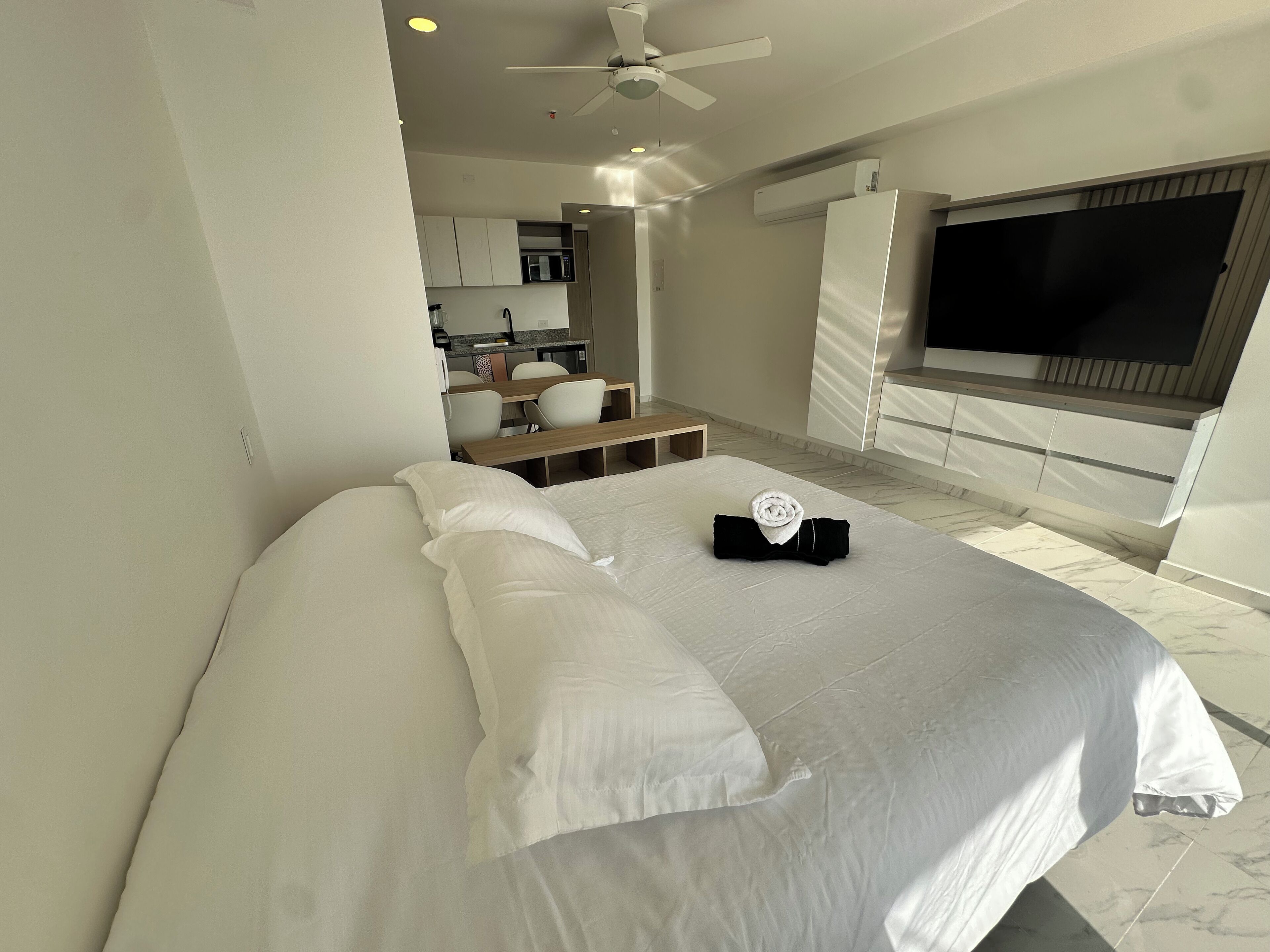 A modern hotel room with a large bed, a TV, a ceiling fan, and a kitchenette in the background.