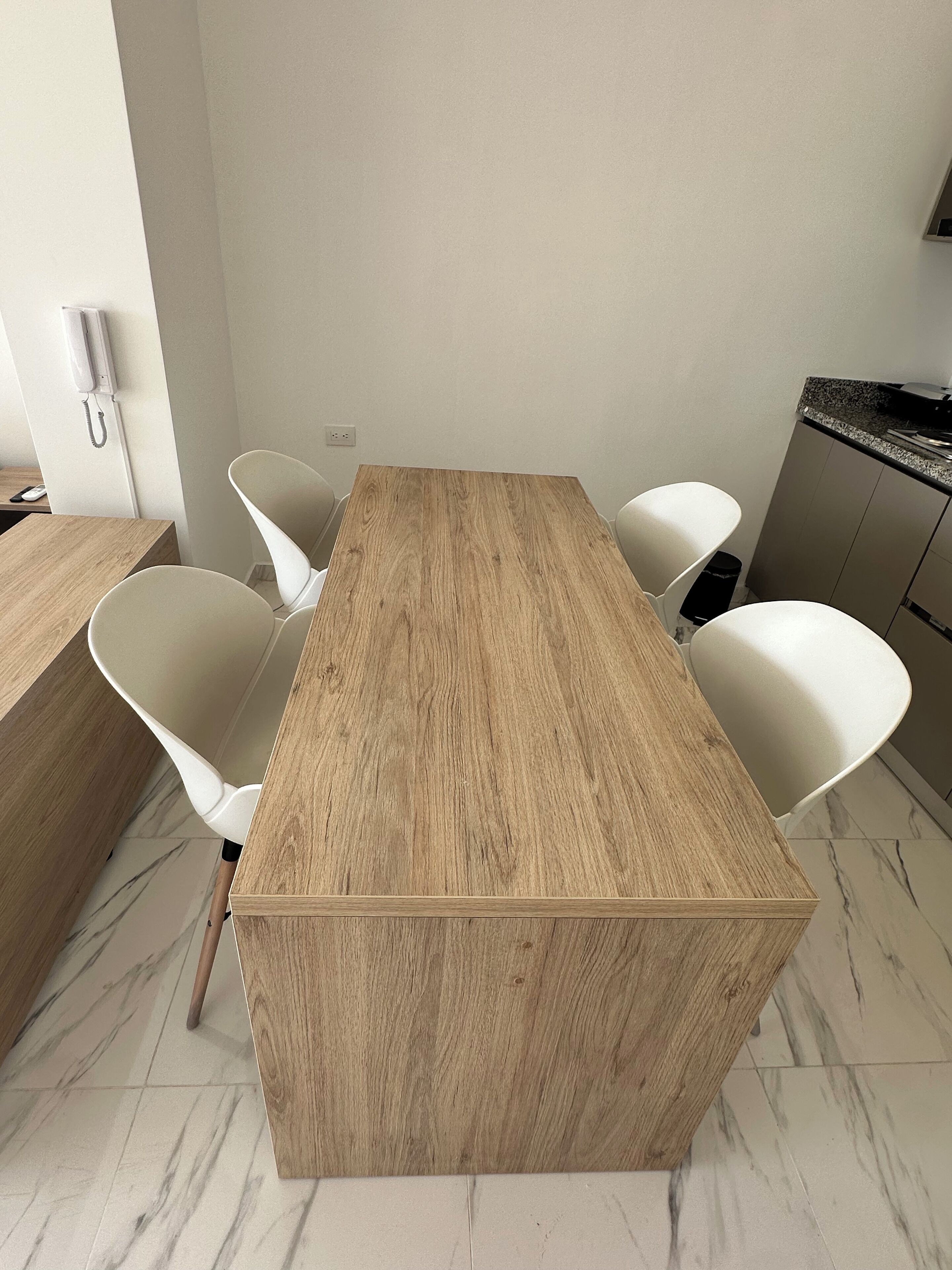 A rectangular wooden table with four white chairs, a white wall, and a marble floor.