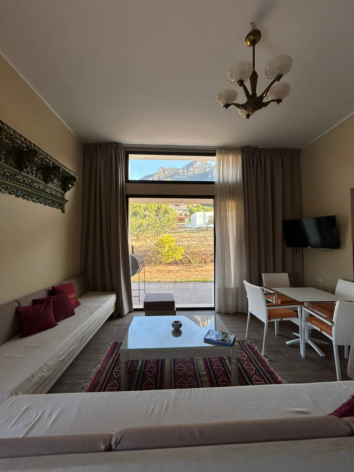 A modern living room with a sofa, a dining table, and a TV. A large window offers a view of a mountainous landscape.