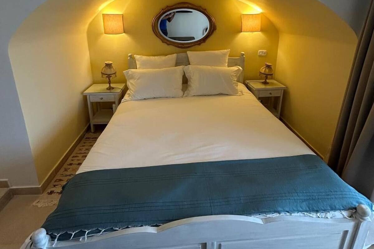 A bed with a white headboard, two bedside tables with lamps, a round mirror above the bed, and a blue throw blanket.