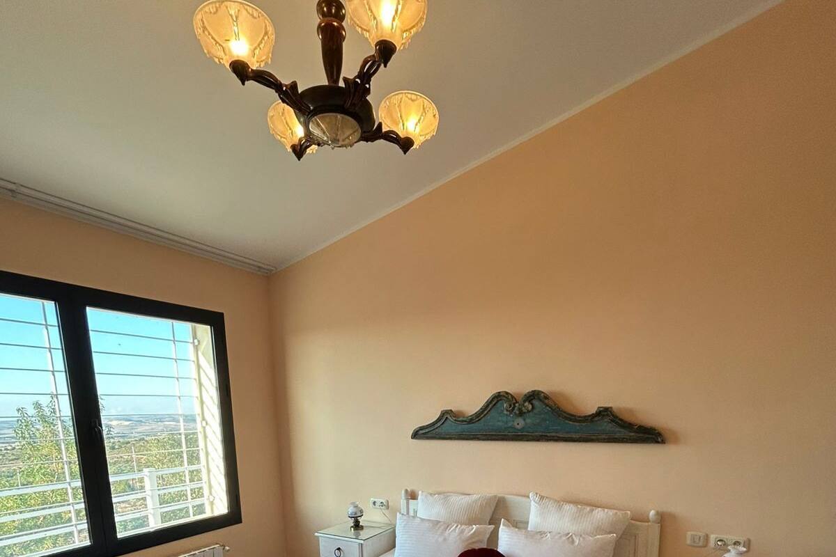 A bedroom with a large bed, two bedside tables, a chandelier, a window with a view, and a red chair.