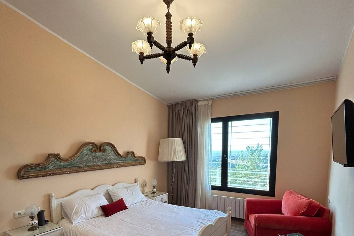 A bedroom with a bed, nightstands, a red sofa, a small table, a TV, and a window with a view.