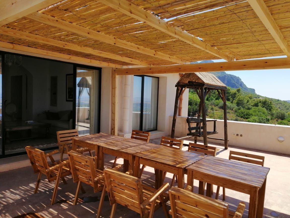 Uma mesa de jantar de madeira com cadeiras num alpendre exterior com vista para uma montanha e vegetação.
