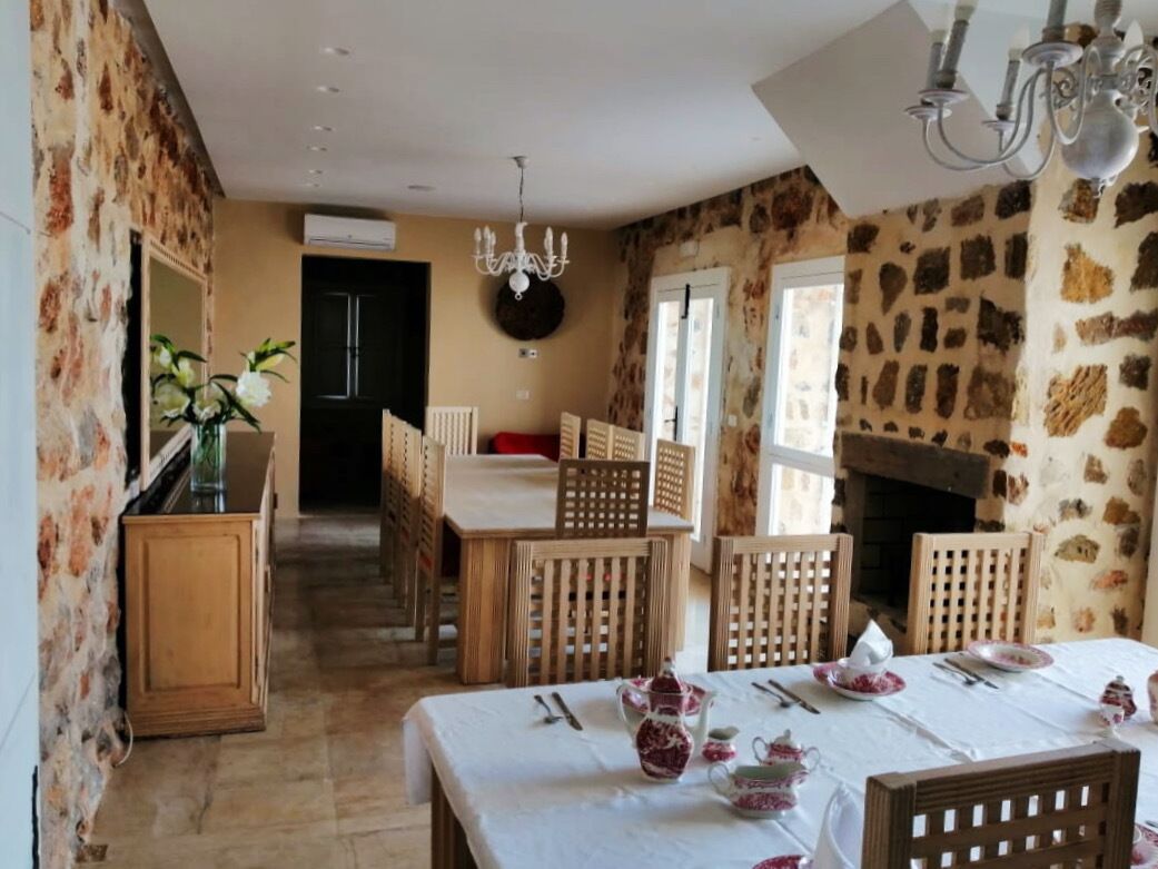 Un coin repas avec une table dressée pour quatre personnes, entourée de chaises en bois. Un lustre est suspendu au plafond, et un mur de pierre est visible sur la droite.