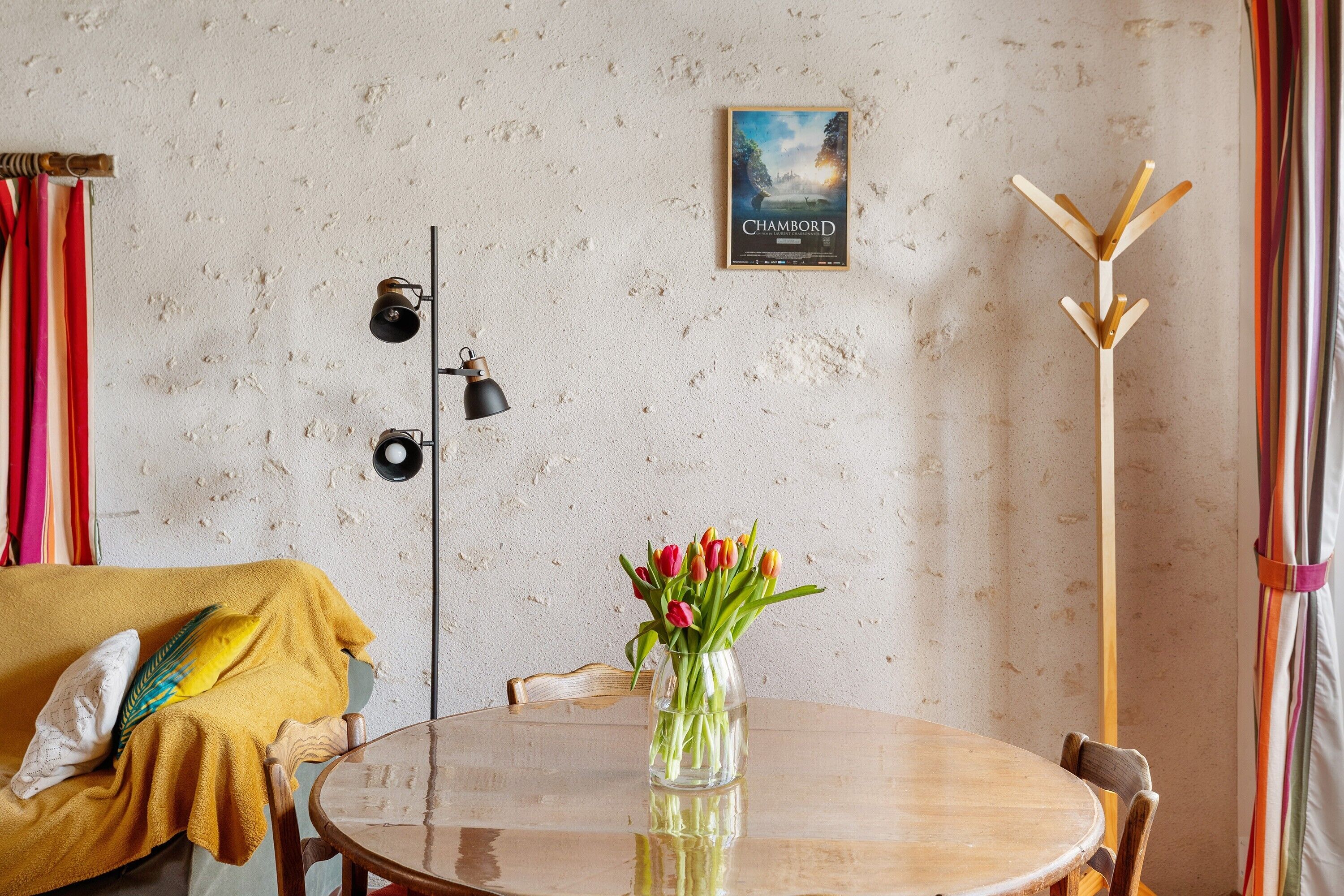 A room with a round wooden table, a vase with tulips, a coat rack, a lamp, a wall clock, and a poster.