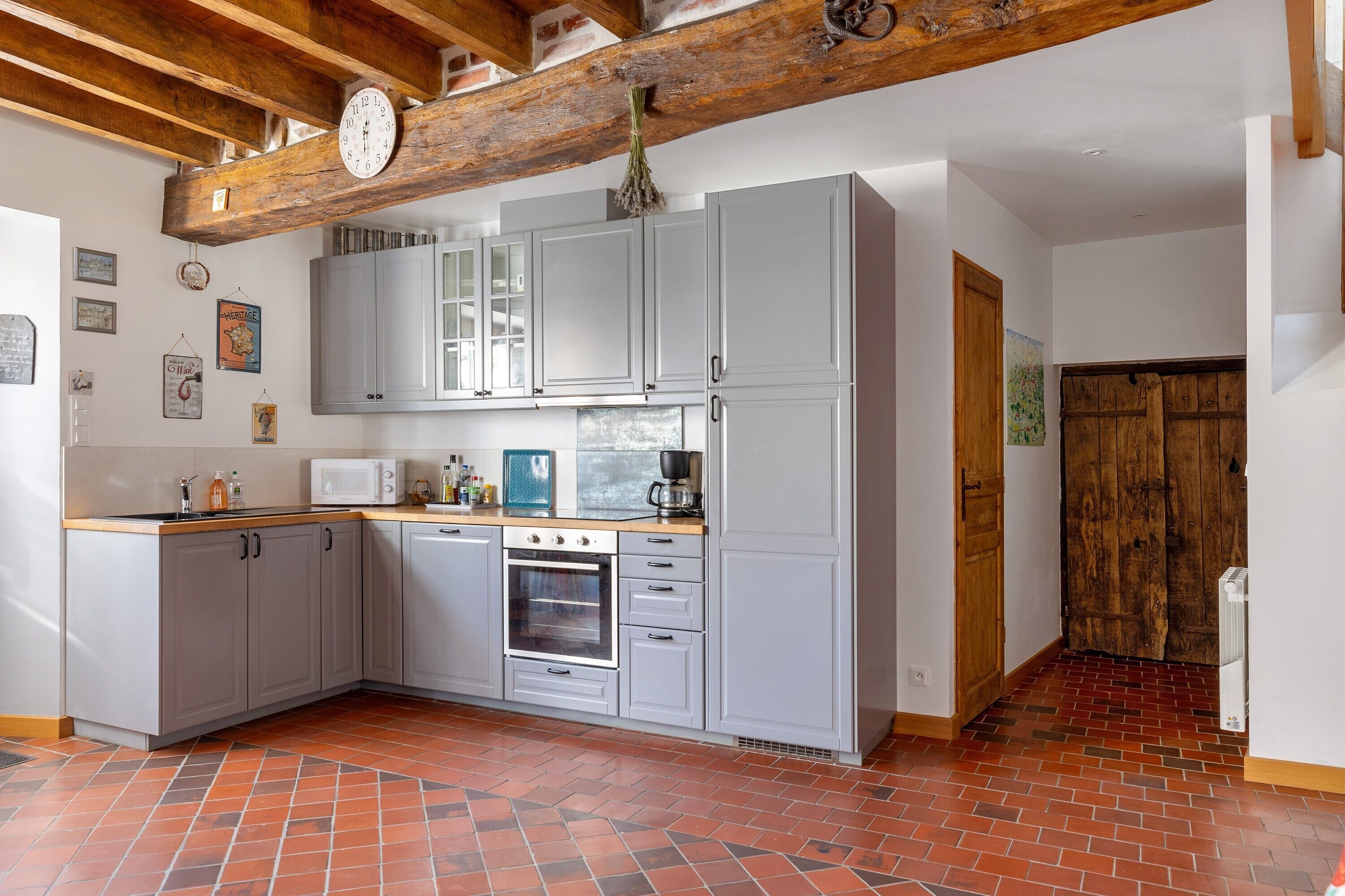 A kitchen with grey cabinets, a wooden ceiling beam, a clock, and a tiled floor.