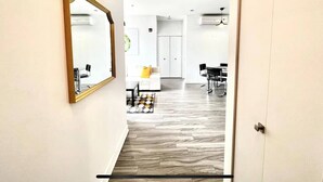 Un pasillo con un espejo, un sofá blanco con un cojín amarillo, un tapete estampado y una zona de comedor con sillas y una mesa.