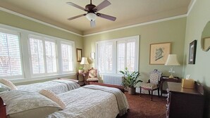 Une chambre avec deux lits, un ventilateur de plafond, de grandes fenêtres équipées de stores, un radiateur, un bureau avec une lampe, une chaise et une plante en pot.