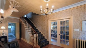 Un escalier avec une rampe en bois et des marches noires, menant à une pièce dont les murs sont recouverts d’un papier peint à motifs et qui est ornée d’un lustre.