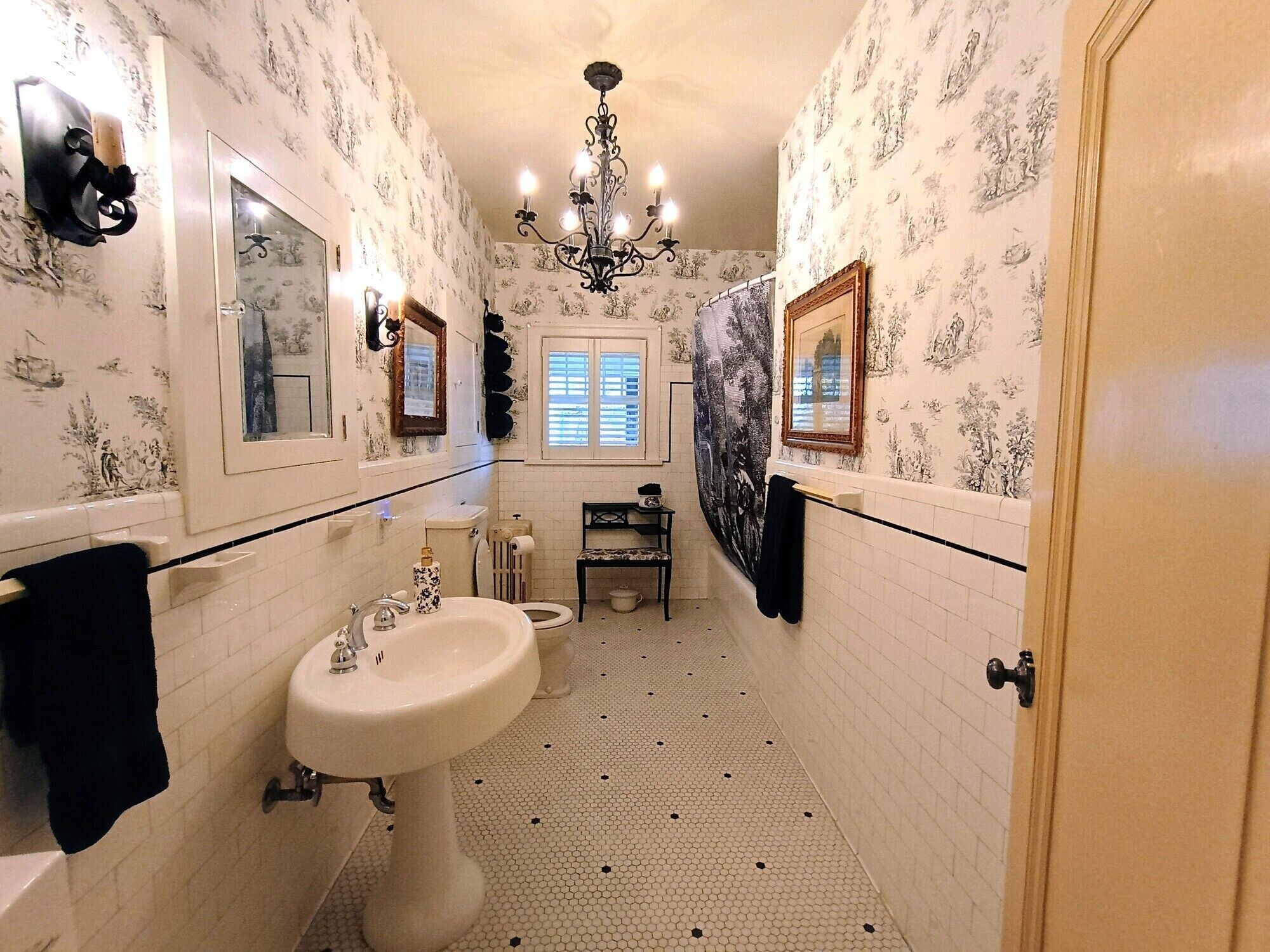 Un baño estrecho con un lavamanos pedestal, dos espejos, una araña y papel tapiz estampado.