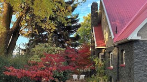 Une maison en pierre avec un toit rouge, entourée d’un jardin aux couleurs automnales et d’un sentier menant à des chaises blanches.