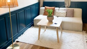 Een gezellige woonkamer met een witte bank, een houten salontafel en een vloerlamp. Er is een raam met gordijnen, een bordje met de WiFi-wachtwoord en een decoratieve wandhanger.
